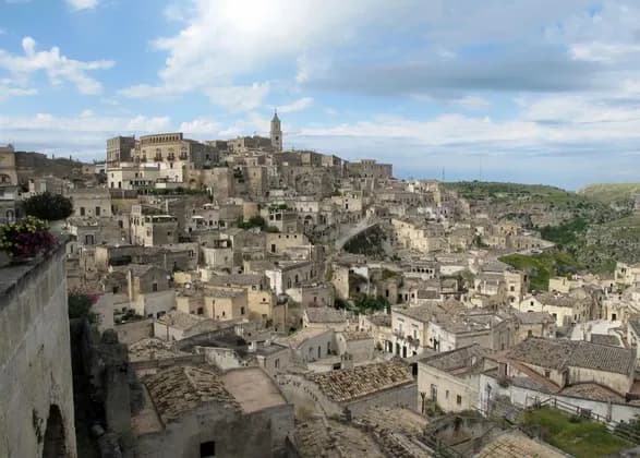 Una vista dall'alto di una città storica con edifici in pietra e tetti in terracotta, costruita sul fianco di una collina sotto un cielo parzialmente nuvoloso.