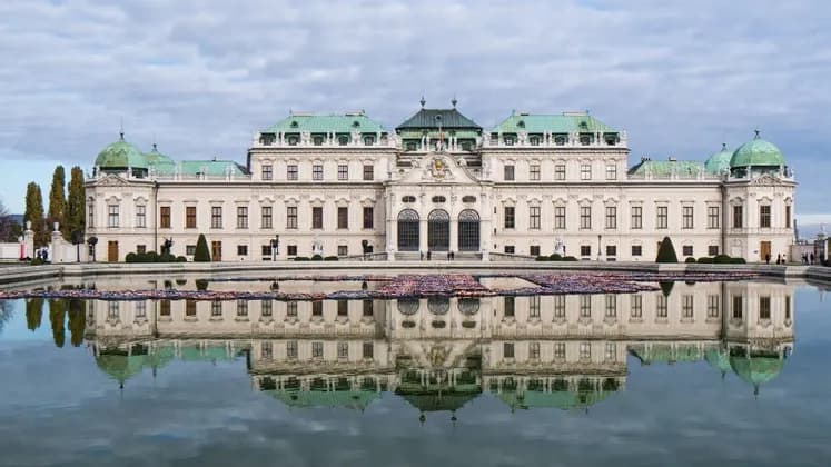 Un palais blanc orné d'un toit vert se reflète parfaitement dans un vaste étang sous un ciel nuageux.