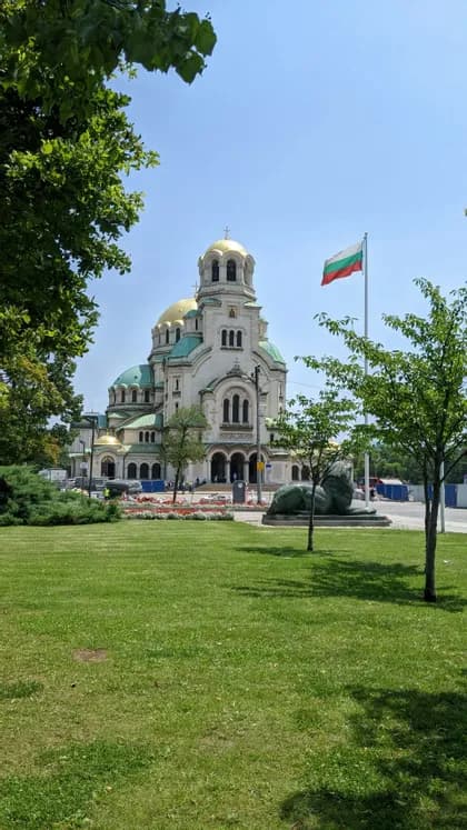 Eine große Kathedrale mit goldenen und grünen Kuppeln und einer bulgarischen Flagge, von einem grünen Park an einem sonnigen Tag aus gesehen.