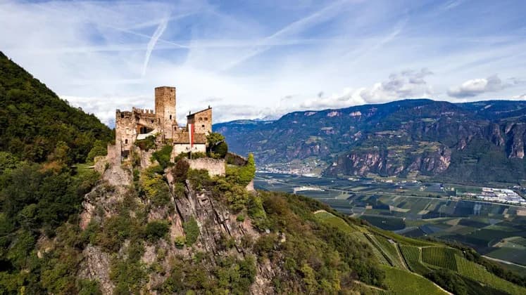 Eine mittelalterliche Steinburg thront auf einer felsigen, baumbestandenen Klippe über einem weiten Tal mit Feldern und Bergen unter blauem Himmel.
