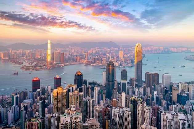 An aerial view of a dense urban cityscape with modern skyscrapers bordering a harbor at sunset.