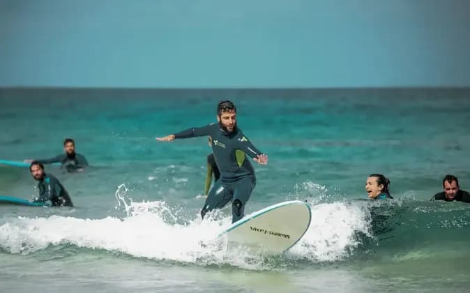 Un uomo del gruppo WeRoad, in muta, sta su una tavola da surf cavalcando un'onda, mentre altri osservano dall'oceano turchese.