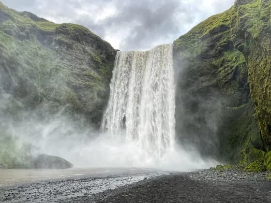 A powerful waterfall cascades down a green, mossy cliff onto a dark pebble shore, creating a thick mist under a cloudy sky.