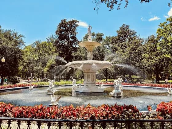 An ornate white tiered fountain with statues spraying water in a public park, with red plants in the foreground and tall trees behind.