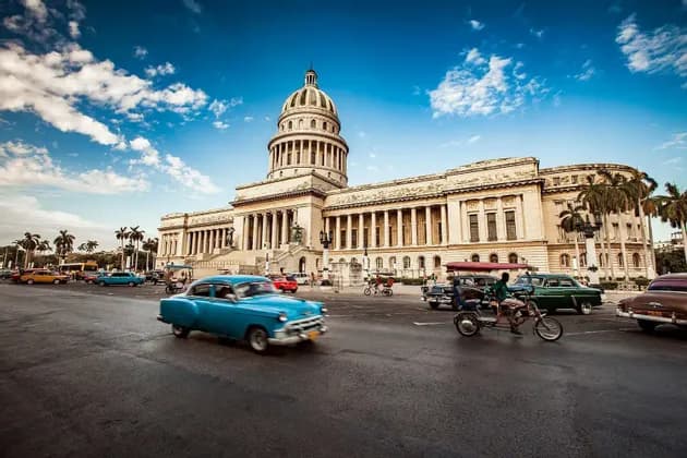 Coches clásicos coloridos circulan por una calle frente a un gran edificio clásico con cúpula bajo un cielo azul.