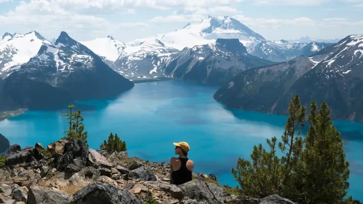 A person in a yellow cap sits on a rocky clifftop, looking out over a bright blue alpine lake surrounded by snow-capped mountains.