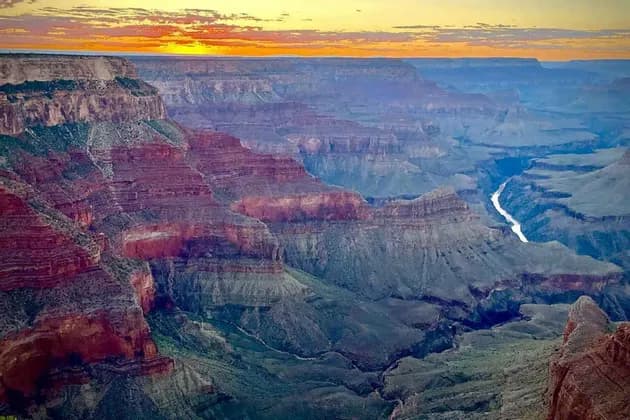 A wide canyon with layered red rock formations and a winding river seen at sunset under an orange sky.