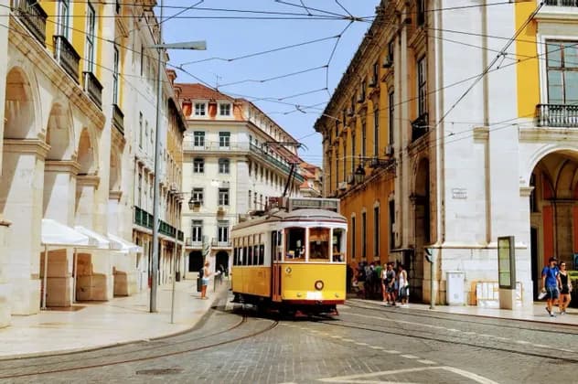 Eine gelbe Straßenbahn fährt auf Gleisen entlang einer Kopfsteinpflasterstraße, umrahmt von historischen Gebäuden mit Bögen unter einem klaren blauen Himmel.