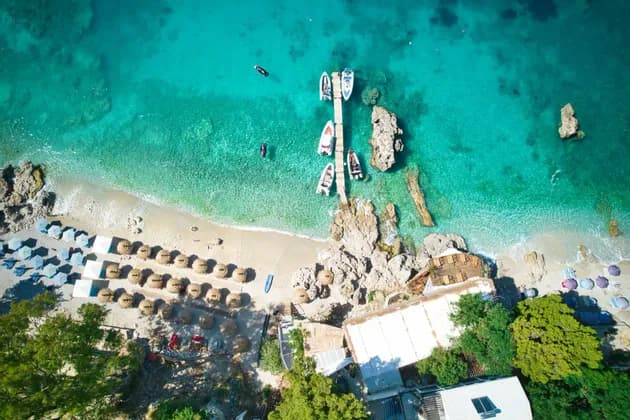 Luftaufnahme eines sonnigen Strandes mit klarem türkisen Wasser, einem Holzsteg mit Booten und Reihen von Strohschirmen.