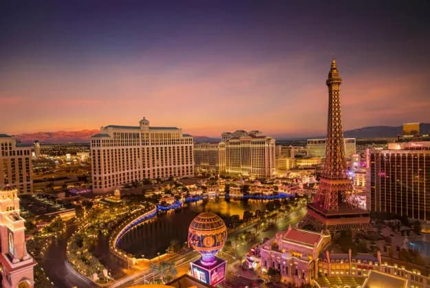 An aerial view of an illuminated city skyline at dusk, featuring a replica of the Eiffel Tower next to large hotels and a lake.