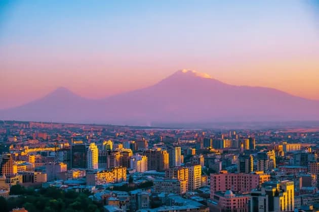 Un paysage urbain au coucher du soleil, avec des bâtiments illuminés et une grande montagne à double sommet à l'horizon sous un ciel aux couleurs vives.
