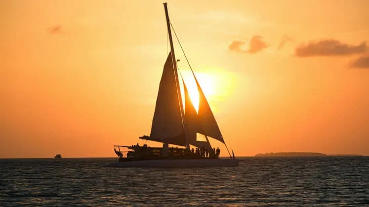 A WeRoad group trip on a catamaran is silhouetted on the ocean against a vibrant orange sunset.