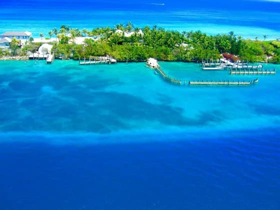 An aerial view of a lush tropical island with buildings and docks, surrounded by turquoise and deep blue ocean.