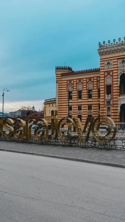 Una grande insegna dorata 'Sarajevo' si erge su un marciapiede davanti a un edificio storico a strisce sotto un cielo nuvoloso.