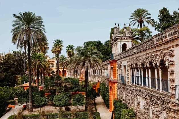 An ornate stone building with a long, arched balcony overlooks a lush garden filled with tall palm trees and pathways.