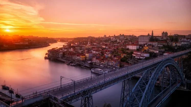 An illuminated arched bridge spans a wide river next to a historic city with red-roofed buildings during an orange sunset.