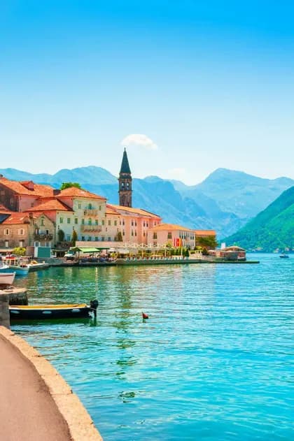 A coastal town with red-tiled roofs and a stone bell tower on the edge of a turquoise bay, with mountains in the distance.