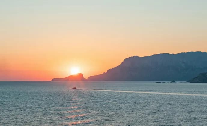 Una barca naviga sul mare durante un tramonto vivace, con isole rocciose stagliate contro il cielo arancione.