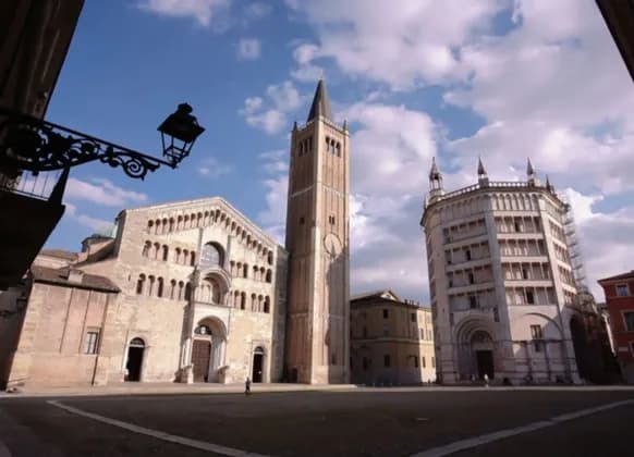 Una storica cattedrale in pietra, campanile e battistero in una grande piazza assolata sotto un cielo blu con nuvole.