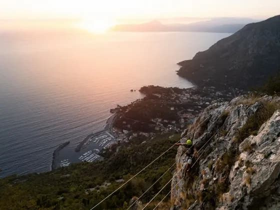 Una persona che scala una via ferrata su una scogliera, con una vista aerea di un porto turistico e della costa al tramonto.