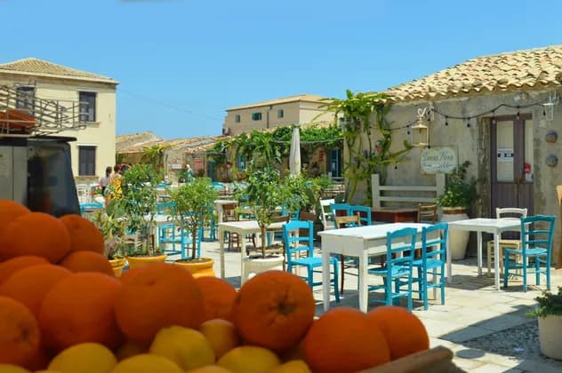 Una cassa di arance fresche in primo piano in un caffè all'aperto soleggiato con sedie blu vivaci e tavoli bianchi.