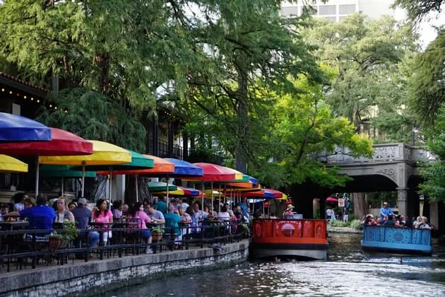 Diners sit at a riverside patio under colorful umbrellas as tour boats float past on the water.