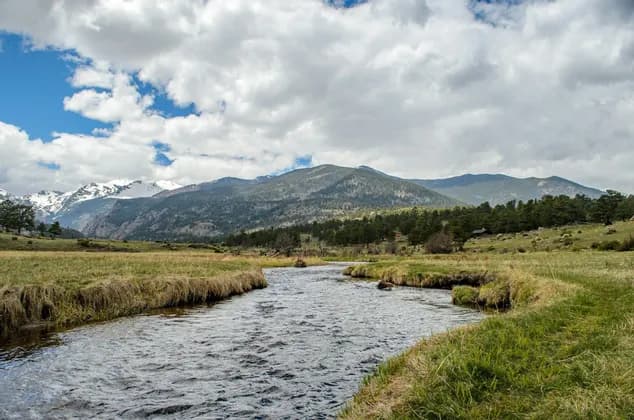 A river flows through a grassy meadow towards a range of snow-capped mountains under a partly cloudy sky.