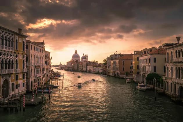 Un ampio canale con barche è fiancheggiato da edifici storici sotto un cielo drammatico al tramonto, con una basilica a cupola in lontananza.