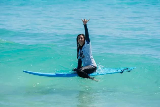 Una donna sorridente in muta siede su una tavola da surf blu in acqua turchese, facendo il segno shaka con la mano.