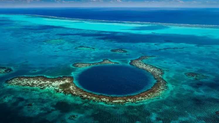 An aerial view of a large, circular marine sinkhole with dark blue water, surrounded by a coral atoll in a turquoise ocean.