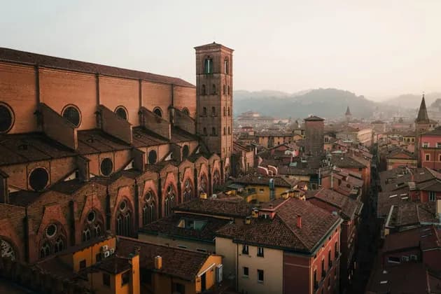 A large brick cathedral with an adjacent bell tower overlooks a historic cityscape with terracotta rooftops under a hazy sky.