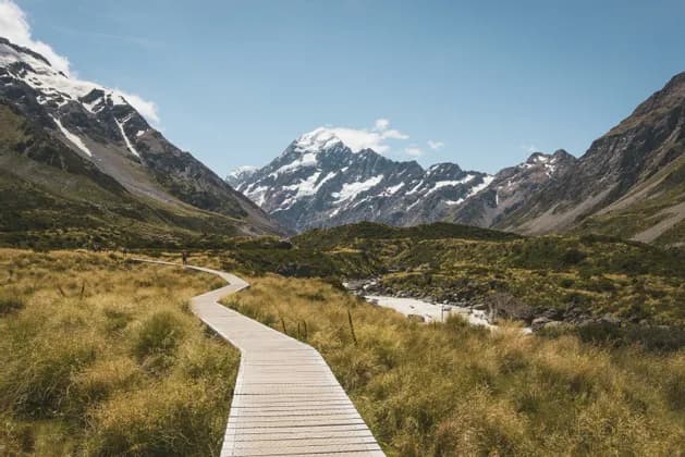 A wooden boardwalk winds through a grassy valley towards towering, snow-capped mountains under a clear blue sky.