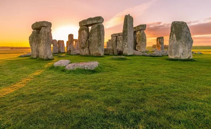 The large standing stones of the Stonehenge monument on a grassy field under a glowing sunrise sky.