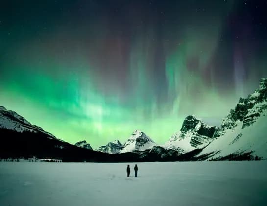 Two people stand on a vast snowy landscape watching the green and purple aurora borealis over snow-capped mountains.