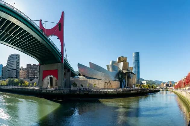 A panoramic view of a modern metallic museum and a red bridge over a river on a sunny day.