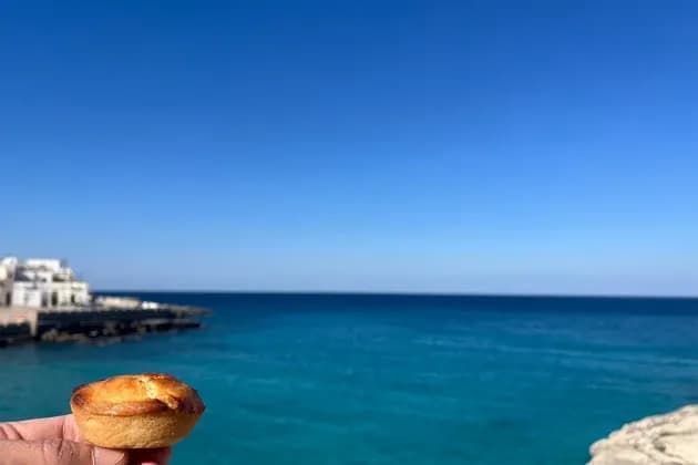 Una mano tiene un piccolo pasticcino davanti a un mare turchese con lontani edifici costieri bianchi sotto un cielo azzurro limpido.