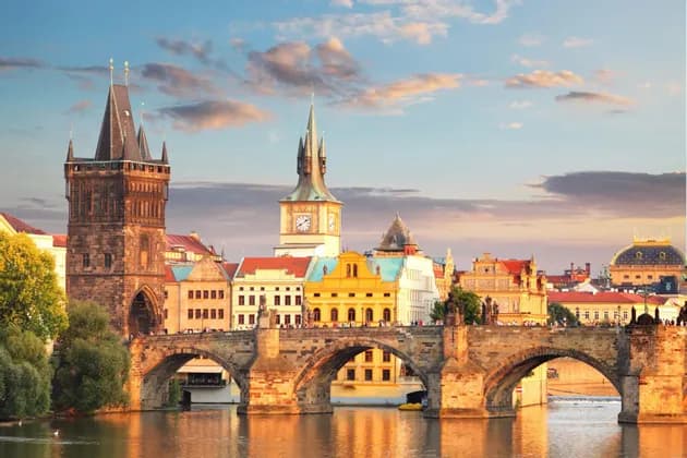 Un pont en arc en pierre historique avec une tour gothique enjambe une rivière devant la ligne d'horizon d'une ville européenne au coucher du soleil.