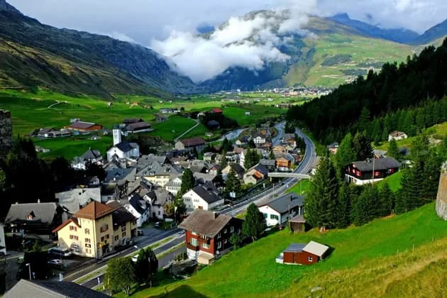 Una vista aerea di un villaggio incastonato in una lussureggiante valle di montagna verde, con una strada che lo attraversa sotto un cielo parzialmente nuvoloso.
