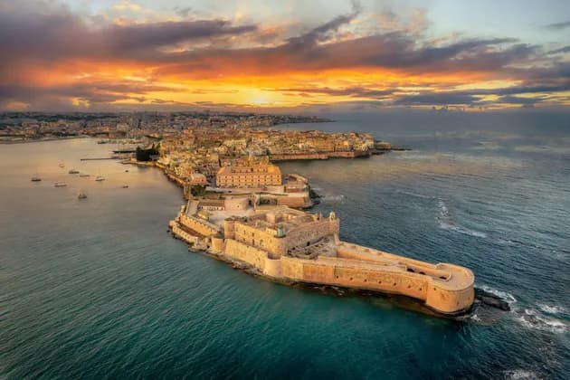 An aerial view of a stone fortress on a peninsula and a coastal city during a colorful sunset over the sea.