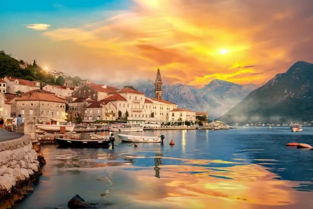 Una storica cittadina costiera con un campanile si affaccia su un porto sereno con barche, incorniciata dalle montagne sotto un tramonto dorato.