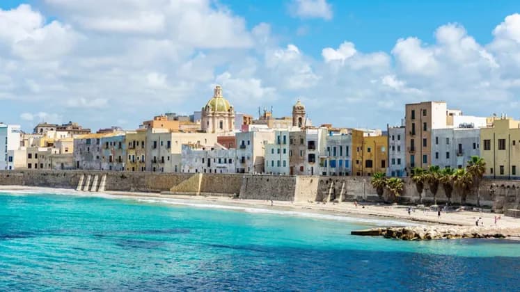 Una città costiera con edifici colorati e una muraglia in pietra si affaccia su una spiaggia sabbiosa con acqua turchese sotto un cielo parzialmente nuvoloso.