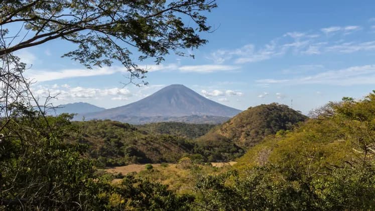 Un grande vulcano a forma di cono si erge su un paesaggio di verdi colline ondulate e foreste, sotto un cielo blu con nuvole leggere.