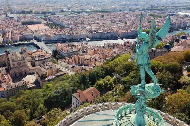 Una statua verde di un angelo alato su una cupola si affaccia su un paesaggio urbano denso con un fiume e edifici dai tetti in terracotta.
