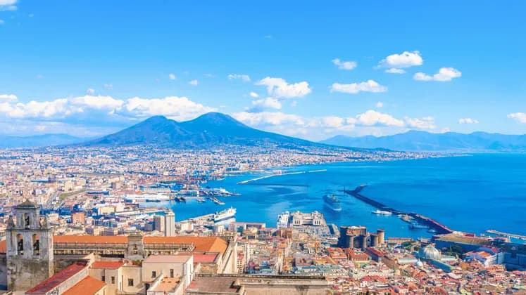Una veduta aerea di una città costiera e della sua baia con un grande vulcano sullo sfondo sotto un cielo azzurro e soleggiato.
