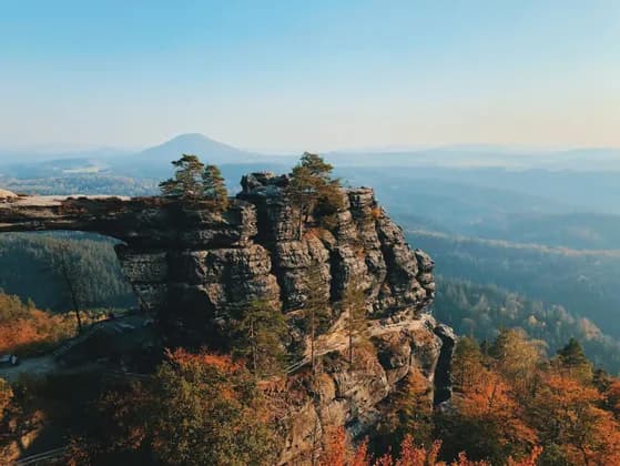 Ein natürlicher Steinbogen auf einer felsigen Klippe überblickt ein Tal mit herbstlich gefärbten Bäumen unter einem klaren Himmel.