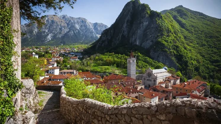 Una vista da un sentiero in pietra che si affaccia su un borgo con tetti in terracotta e una chiesa, incastonato in una lussureggiante valle montana.