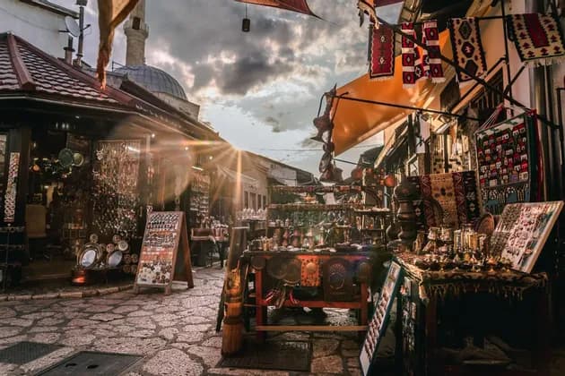 Un callejón empedrado bañado por el sol está bordeado de puestos de mercado que venden artesanías tradicionales, con una cúpula visible contra un atardecer nublado.