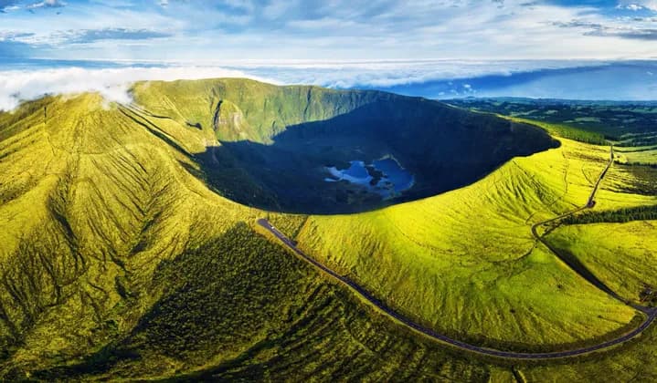 Vista aerea di un grande cratere vulcanico verde, con un lago alla base e una strada che serpeggia lungo il suo bordo, sotto un cielo parzialmente nuvoloso.