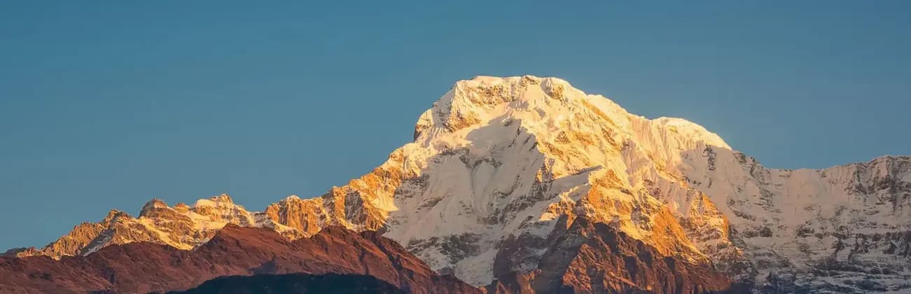 Un imposant sommet montagneux enneigé, illuminé par les rayons dorés du soleil, se découpe sur un ciel bleu clair.