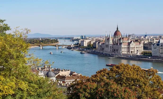 Una veduta sopraelevata e soleggiata di un grande edificio sontuoso sulla riva di un fiume, con barche, un ponte e una città sullo sfondo.